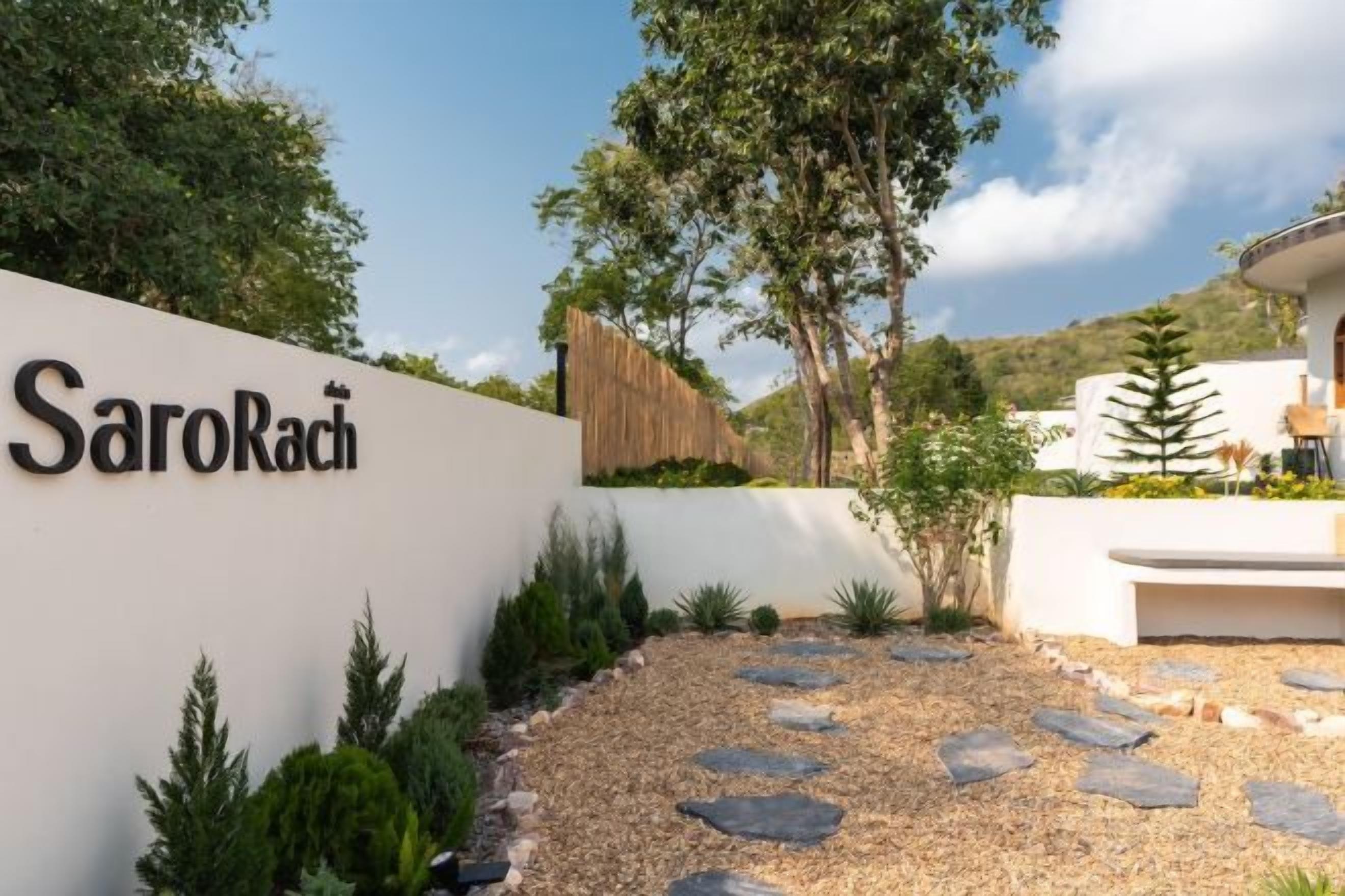 A modern building entrance with a white wall displaying the name 'SaroRach' and a gravel pathway leading to the entrance.