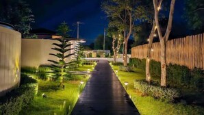 Un chemin nocturne bordé d’arbres et éclairé par des lampadaires, menant à un bâtiment au design moderne.