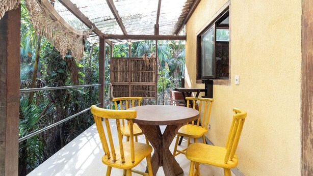Un espacio de comedor al aire libre cubierto, con una mesa redonda y sillas amarillas, rodeado de abundante vegetación.