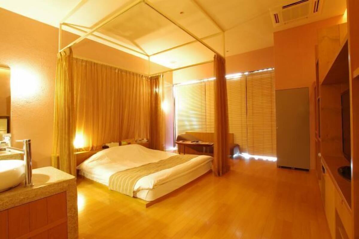A hotel room with a bed, a bedside table, a TV, a bathroom with a sink, and a window with blinds.