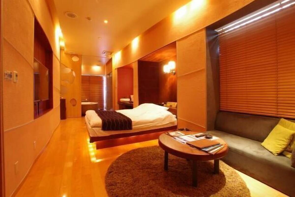 A modern hotel room with a bed, a sofa, a round coffee table, and a large window with blinds.