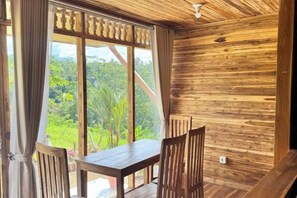 Une pièce en bois avec une table et des chaises, de grandes fenêtres avec des rideaux et une vue sur la verdure à l’extérieur.