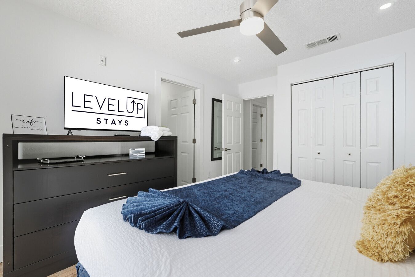 Une chambre d’hôtel avec un lit, une commode sombre, un panneau indiquant « LevelUp Stays », et un ventilateur de plafond.