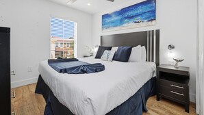 Une chambre moderne avec un grand lit, un ventilateur de plafond, un tableau, une table de chevet et une fenêtre avec des stores.