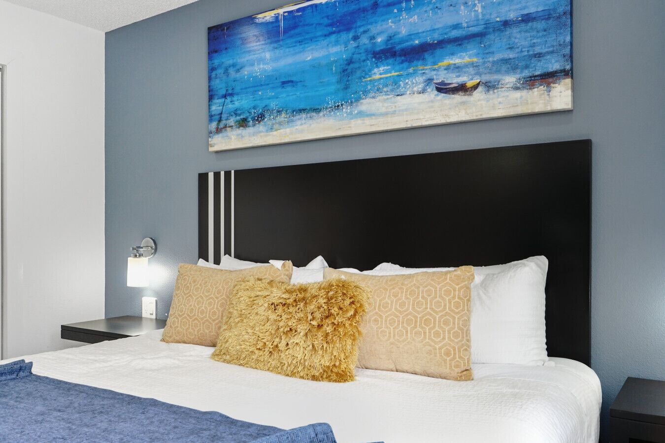 A modern bedroom with a large bed, a headboard with vertical stripes, and a painting of a blue seascape.