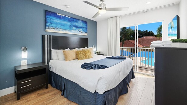 Une chambre moderne avec un grand lit, un ventilateur de plafond et une vue sur une piscine et un immeuble.