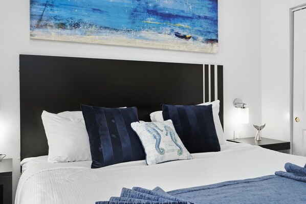 Una habitación de hotel con una cama grande, ropa de cama blanca y una almohada con forma de caballito de mar azul. Hay un cuadro de un océano azul en la pared sobre la cama.