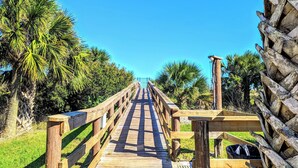 A wooden boardwalk leading to a distant structure, surrounded by palm trees and greenery.