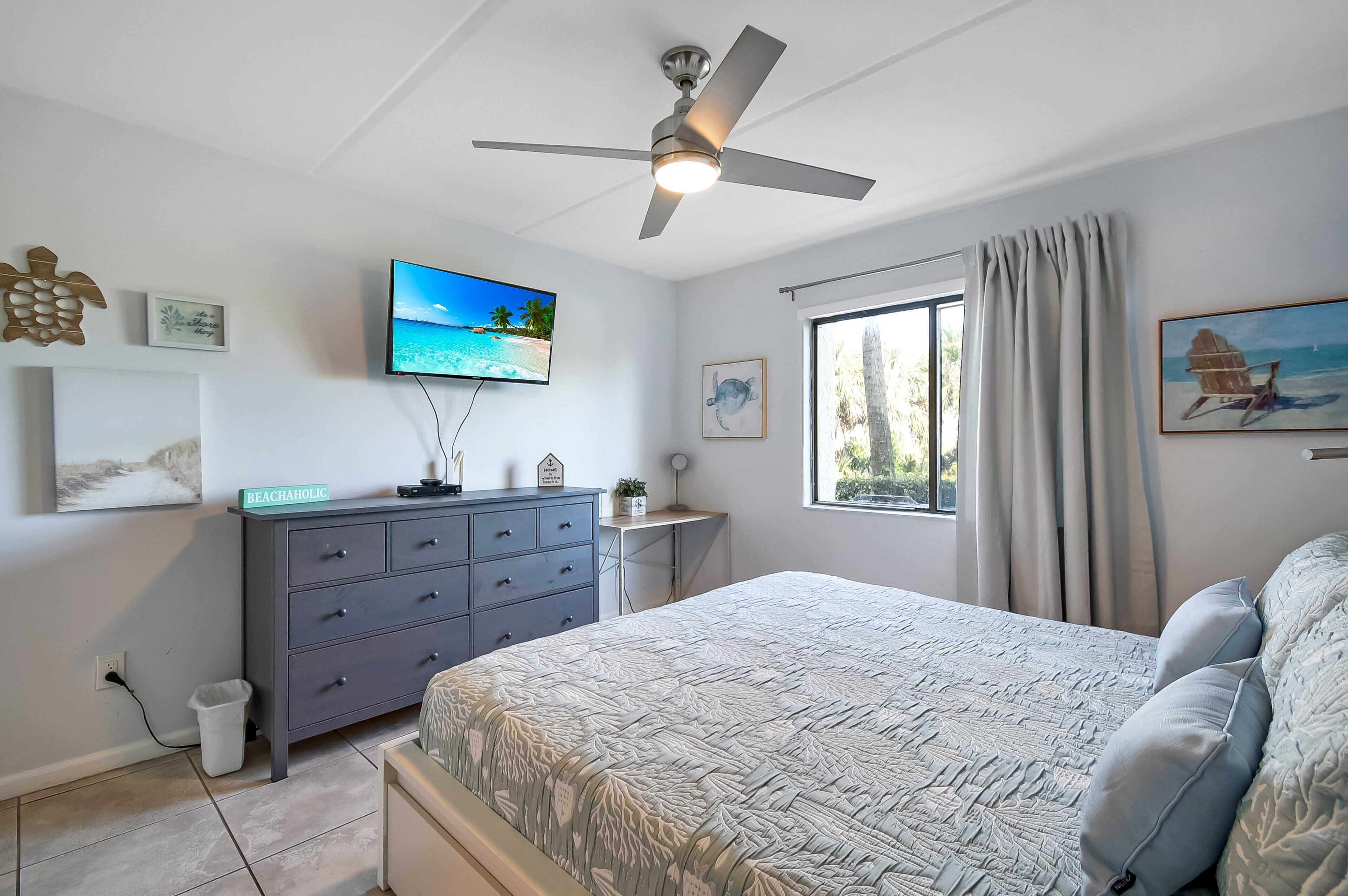 Ein Schlafzimmer mit einem Bett, einem Schreibtisch, einem Deckenventilator, einem Fernseher und einem Fenster mit Vorhängen.
