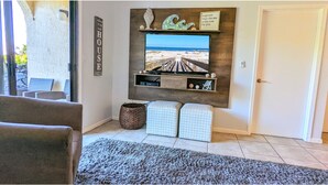 A living room with a large TV mounted on a wooden wall, a beach view, and a cozy seating area with a rug.