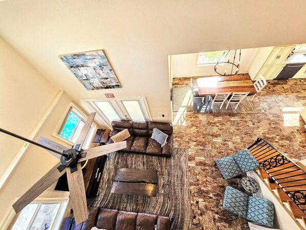 A spacious living area with a brown leather couch, a wooden coffee table, and patterned ottomans. A ceiling fan and a painting are visible.