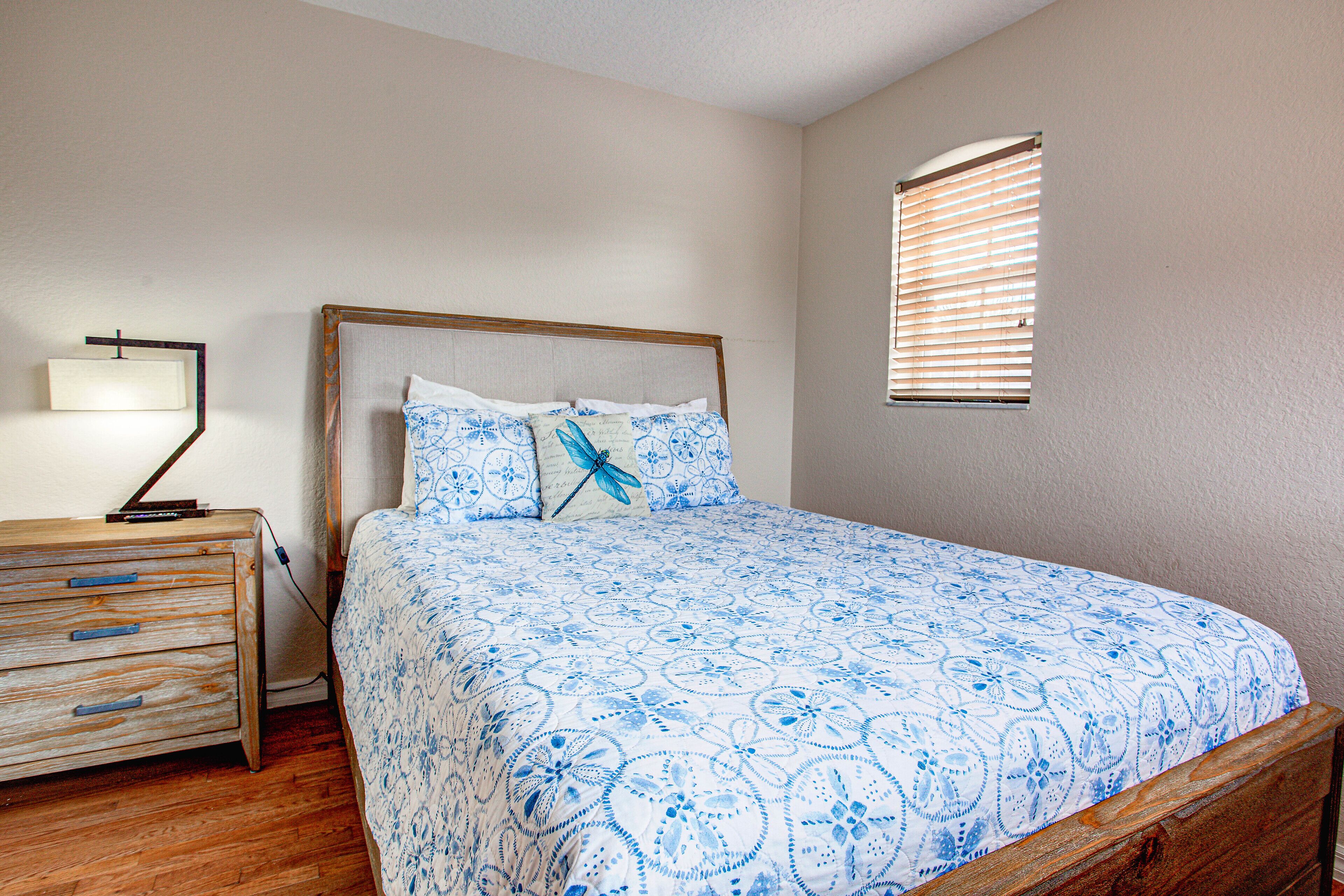 A bedroom with a wooden bed frame, a blue floral bedspread, a nightstand with a lamp, and a window with blinds.
