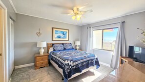 Un dormitorio con cama, dos mesitas de noche, ventilador de techo, ventana con cortinas y un mueble para televisión.