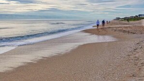 Una playa con gente caminando por la orilla, un mar tranquilo y casas al fondo.
