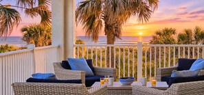 A balcony with wicker furniture, a table, and a view of the sunset over the ocean.