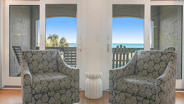 A room with two patterned armchairs facing a sliding glass door leading to a balcony with a view of the ocean.