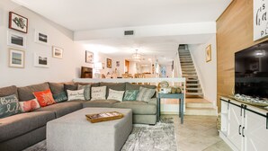 A living room with a sectional sofa, ottoman, and a flat-screen TV. There is a staircase leading to an upper level.