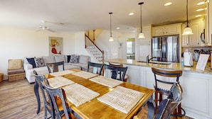 A spacious living area with a wooden dining table, chairs, and a sofa. A kitchen with a stainless steel refrigerator and a countertop is visible in the background. - Oceanfront townhome with a furnished balcony, outdoor pool, & tennis (Fernandina Beach)