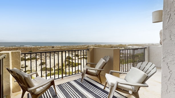 A balcony with two Adirondack chairs, a striped rug, and a view of the beach and ocean. - Oceanfront townhome with a furnished balcony, outdoor pool, & tennis (Fernandina Beach)