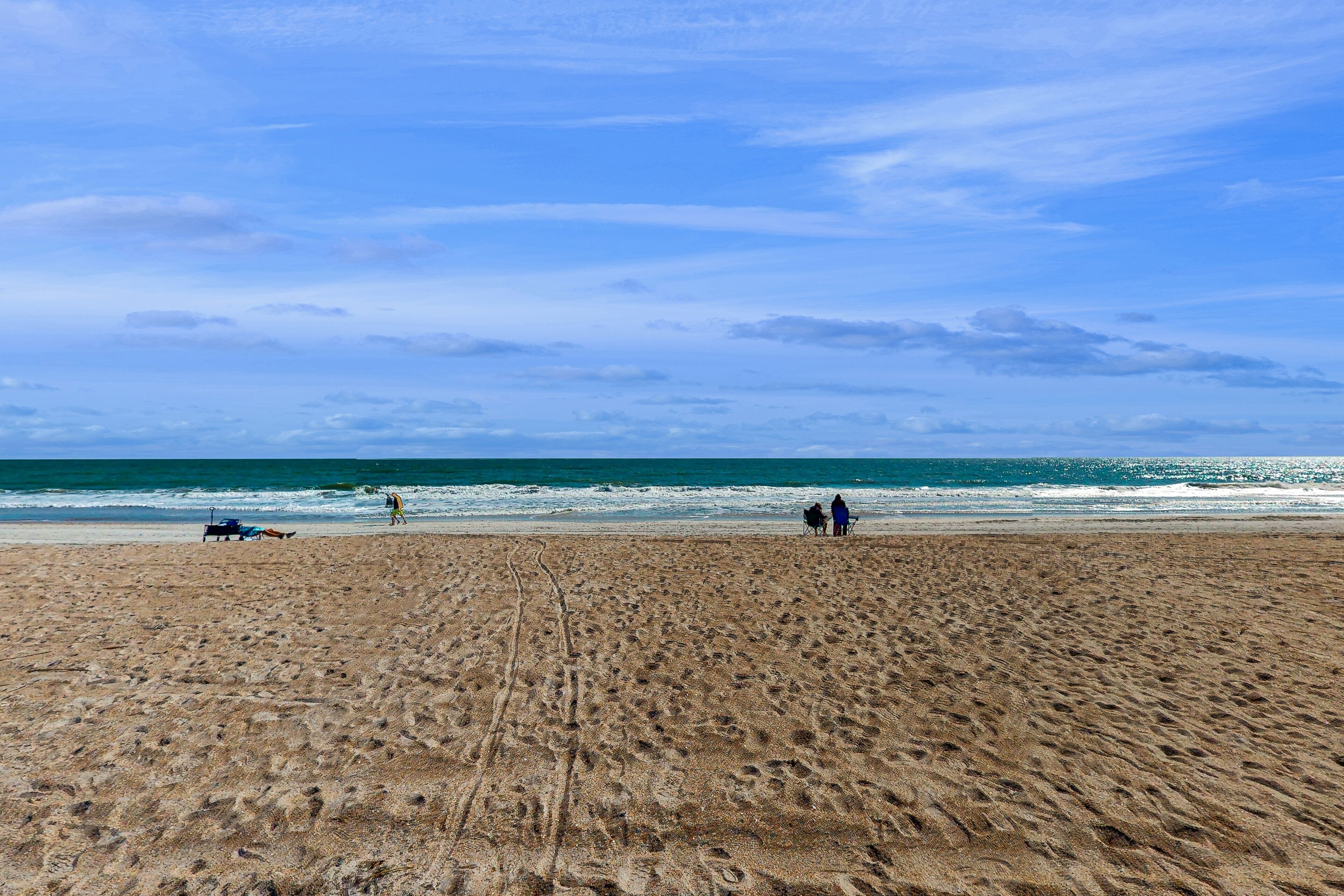 Une vaste plage de sable fin, parsemée d’empreintes menant à l’eau, un ciel dégagé et quelques personnes visibles au loin.