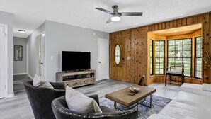 Un salon moderne doté d’un téléviseur à écran plat, de fauteuils en cuir foncé, d’une table basse en bois et d’une grande fenêtre donnant sur un paysage verdoyant.