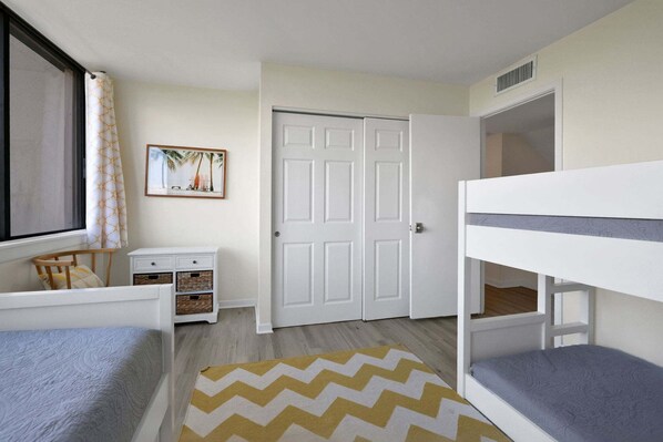 Quarto com beliches, uma escrivaninha, uma cadeira, janela com cortinas, uma porta e um quadro na parede.