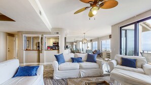 Un salon spacieux avec des canapés blancs, des coussins bleus, une table basse en bois et un ventilateur de plafond.