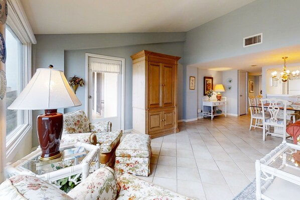 A living room with floral-patterned sofas, a wooden cabinet, and a dining area with white chairs and a chandelier.