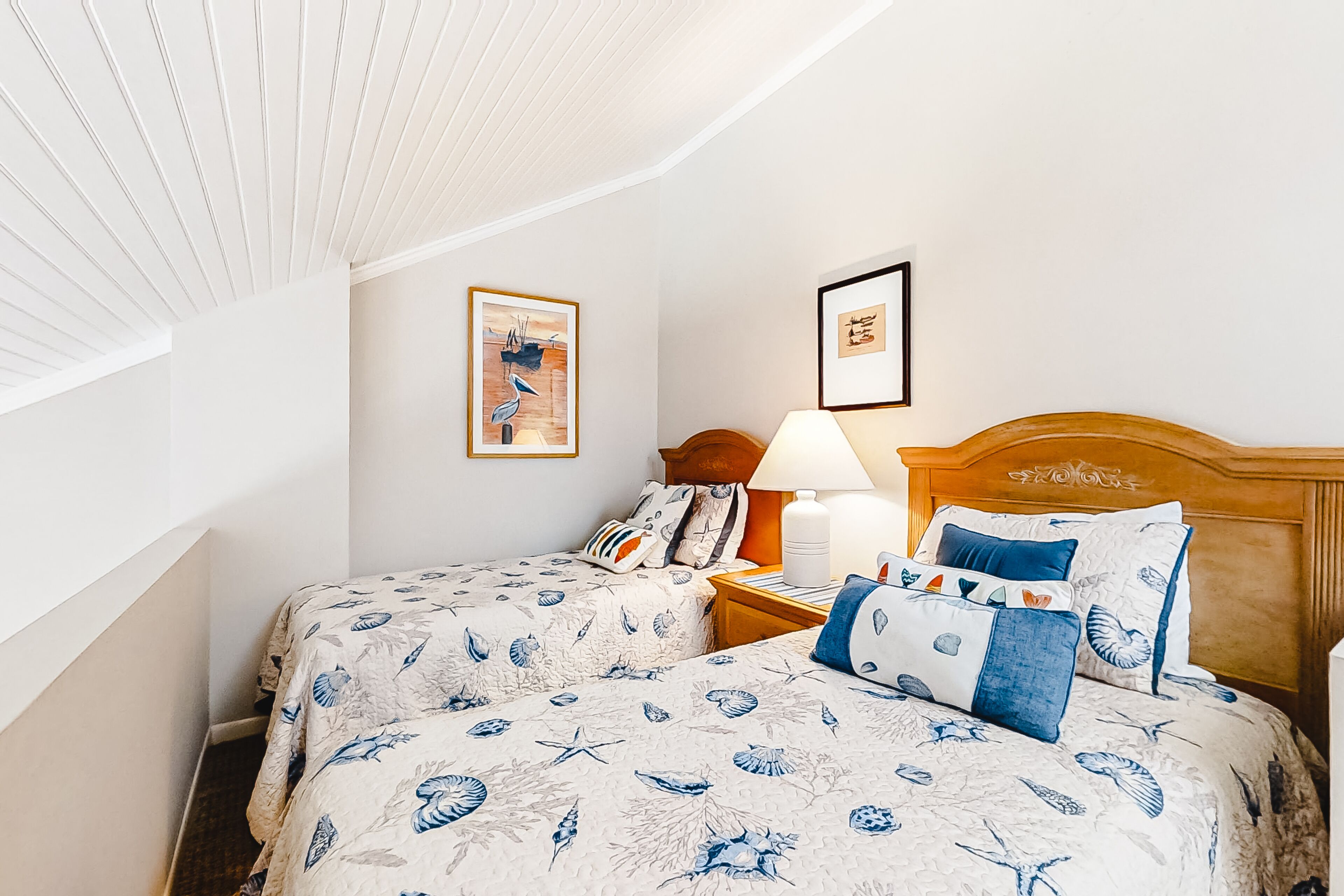 Une chambre avec deux lits, une tête de lit en bois, une table de chevet avec une lampe et des œuvres d’art encadrées accrochées au mur.