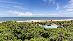 A resort with a swimming pool surrounded by lush greenery, overlooking a beach and the ocean. - Oceanfront condo with balcony, water views, AC & outdoor pool (Fernandina Beach)