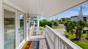 Een balkon met een witte reling, een overdekt terras met een plafondventilator en uitzicht op een woonstraat met huizen en palmbomen.