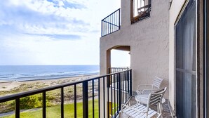 Un balcón con vista al océano, un pequeño espacio de estar al aire libre y un edificio con fachada texturizada.