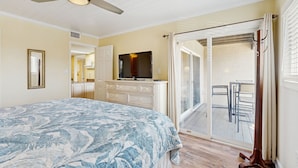 Une chambre à coucher comprenant un lit, un ventilateur de plafond, un téléviseur, une porte-fenêtre coulissante donnant sur un balcon et une commode.