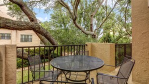 A patio with a round table and four chairs, surrounded by a metal railing, overlooking a garden with trees and a building in the background. - Newly Renovated Summer Beach Village Home with Patio, WiFi & Shared Pool (Fernandina Beach)