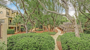 A courtyard with a paved walkway, surrounded by well-maintained hedges and trees, leading to a wooden bridge. - Newly Renovated Summer Beach Village Home with Patio, WiFi & Shared Pool (Fernandina Beach)