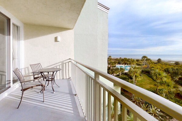 A balcony with a table and chairs, overlooking a pool and lush greenery. - Oceanfront condo with large balcony, pool, central AC & views (Fernandina Beach)