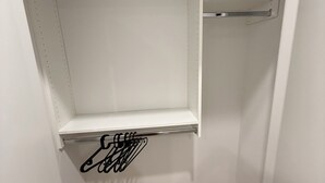 Een kleine, afgesloten kast met witte wanden en een houten vloer. Er zitten twee open planken en een kledingstang in.