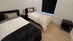 Een slaapkamer met twee bedden, een nachtkastje met lamp, een raam met jaloezieën en een klein tafeltje.