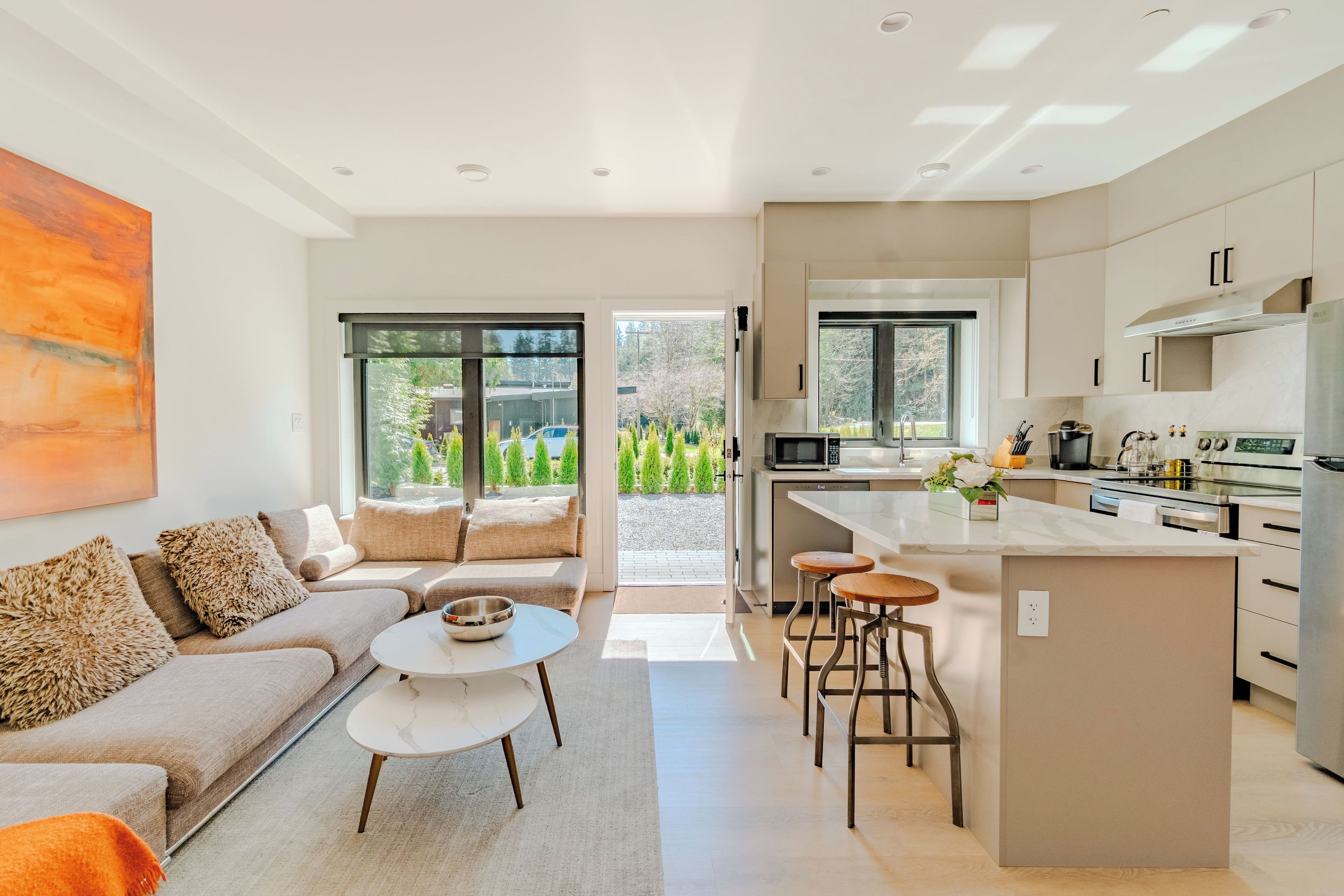 A modern kitchen with a central island, a dining area with a sofa and coffee table, and a view through the glass doors.
