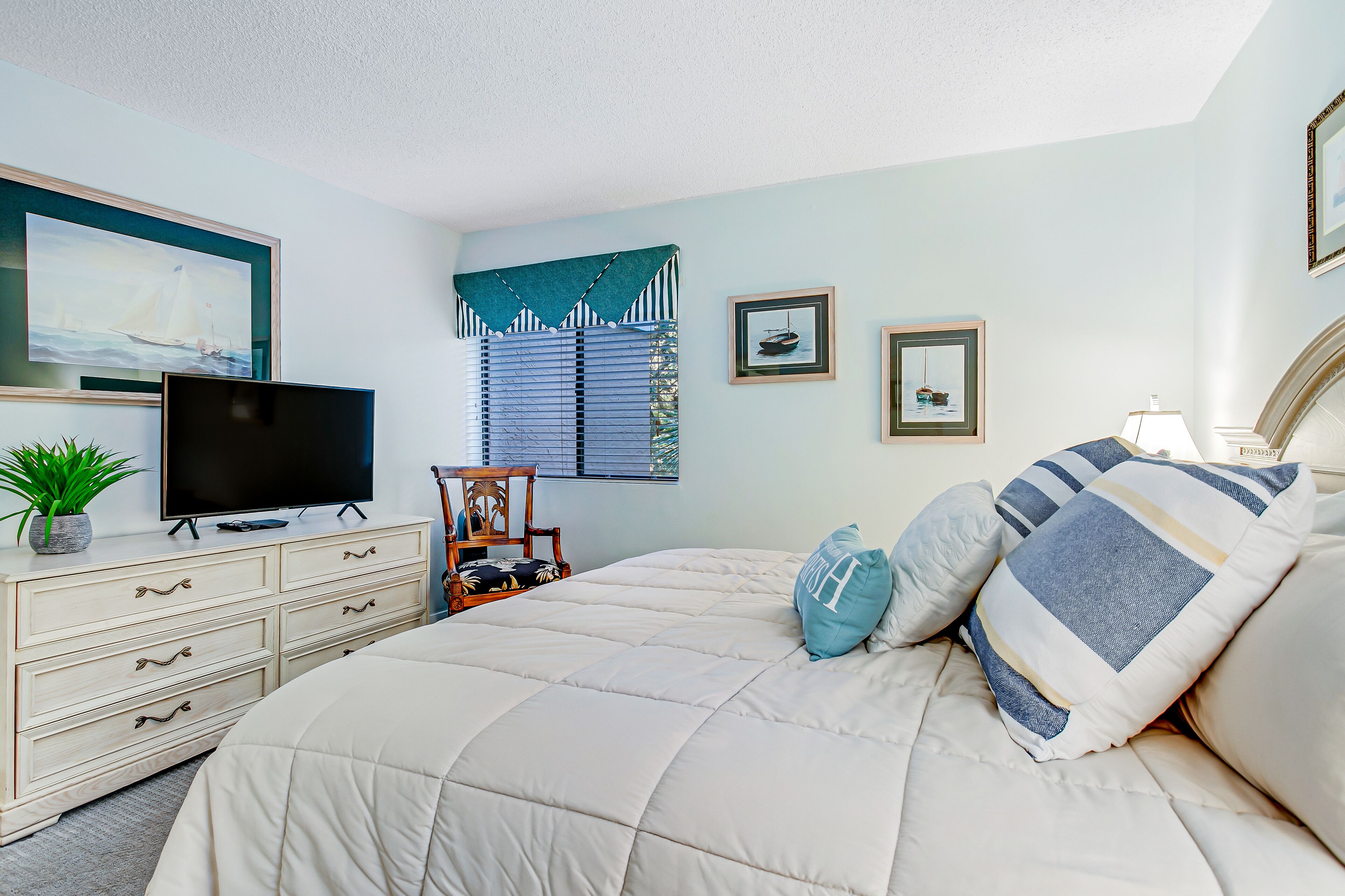 A bedroom with a bed, a TV on a dresser, a window with blinds, and framed pictures on the wall.