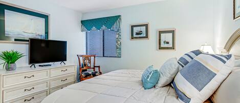 A bedroom with a bed, a TV on a dresser, a window with blinds, and framed pictures on the wall.