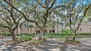 A tree-lined driveway leading to a building with multiple balconies and a sign indicating a parking area.