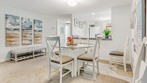 A dining area with a glass table, white chairs, and a bench. There are paintings on the wall and a plant on the counter.