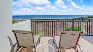 A balcony with two wicker chairs and a glass table, overlooking a beach with a clear sky and distant ocean.