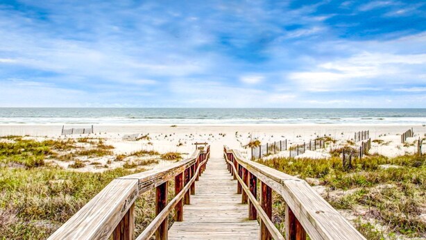 A wooden boardwalk leading to a beach with sand dunes and a fence.