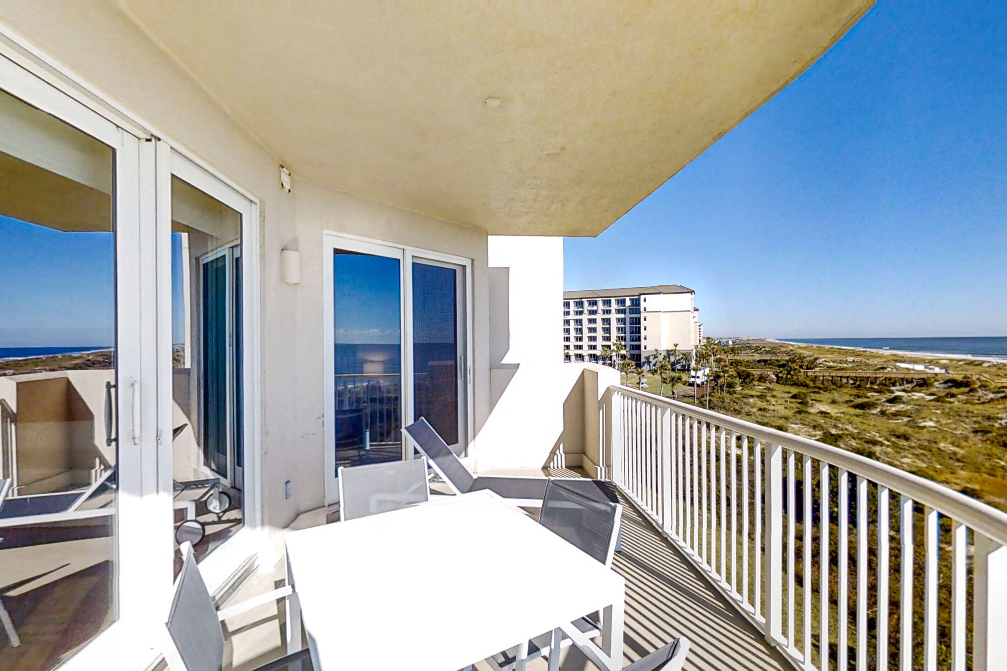 A balcony with a table and chairs, a view of the beach, and a building in the background.