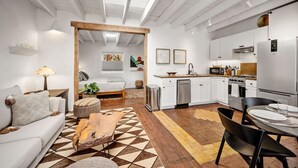 A modern living room with a sofa, coffee table, and a patterned rug. A dining area with a table and chairs is adjacent to the kitchen, which features white cabinets and stainless steel appliances. - El Vaso Lleno - Fantastic Location, Historic Home (Santa Fe)