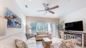 Un salon avec un canapé beige, un fauteuil à motifs et une table basse. On y trouve un ventilateur de plafond, un téléviseur posé sur un meuble en bois et un tableau accroché au mur.