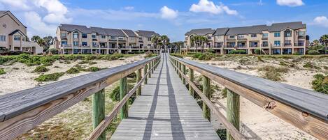 A wooden boardwalk leading to a beachfront property with multiple-story buildings, some with balconies.