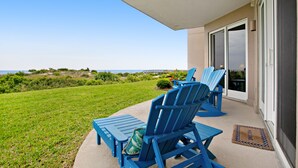 A patio with blue Adirondack chairs and a small table, overlooking a grassy area and the ocean. - Luxe, Updated, Oceanfront Condo with Ground-Floor Patio, WiFi & Shared Pool (Fernandina Beach)